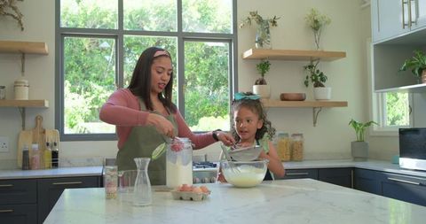 Mother and daughter baking in bright modern kitchen, scooping flour and whisking batter