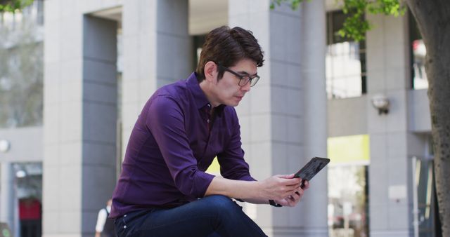 The man is wearing casual attire and seated near a modern office building. He looks relaxed while focusing on his smartphone. Useful for topics about business, technology, communication, and urban lifestyle.