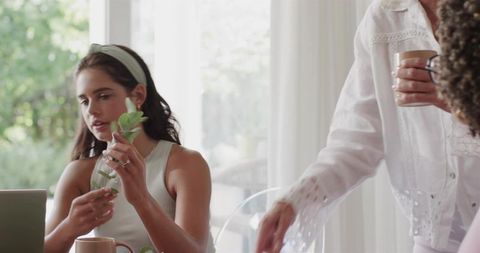 Young woman examining plant cutting while working on laptop as friend reaches for mug