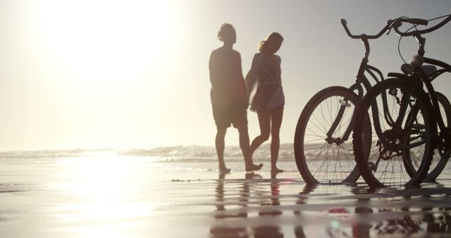A young Caucasian couple enjoys a romantic walk along the beach at sunset, with bicycles parked in the foreground, with copy space. Their silhouettes against the shimmering ocean create a serene and intimate atmosphere.