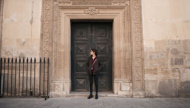 Woman standing against ornate wooden doorway wearing padded jacket and crossbody bag