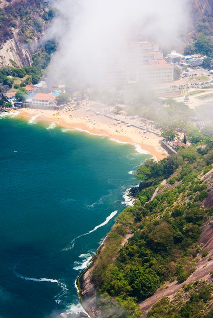 The image showcases a beautiful aerial view of a coastline with a sandy beach flanked by lush green foliage and cliffs, during a misty morning. This photograph can be used in travel brochures, articles about tropical destinations, environmental campaigns, nature blogs, or social media posts promoting beach vacations.