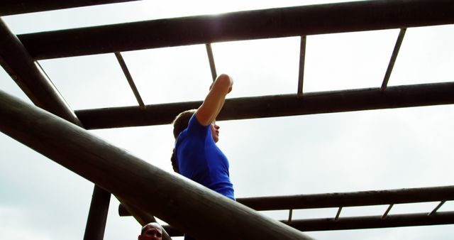 A woman in a blue top confidently navigates monkey bars on an outdoor obstacle course. This image is perfect for illustrating themes related to fitness, determination, outdoor training, physical challenges, and personal growth. It can be used in advertisements, motivational materials, fitness blogs, and promotional content for outdoor activity programs.