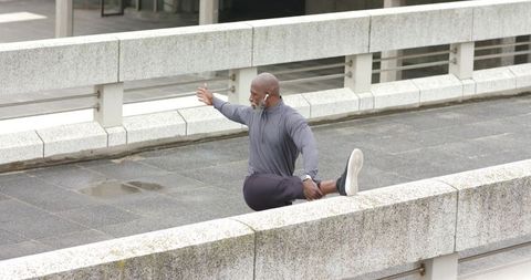 Mature man stretching on urban bridge barrier wearing earbuds, face covering for wellness
