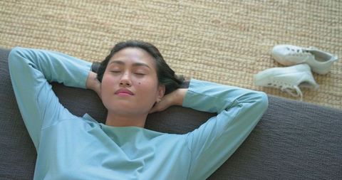 Mid adult asian woman reclining on sofa, relaxing in minimalist living room with sneakers