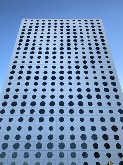 This image of a modern high-rise building with circular windows provides a striking example of contemporary architecture. The building stands tall against a clear blue sky, creating an impressive visual contrast. Ideal for use in architectural design presentations, urban development proposals, or promotional materials for real estate. The repetitive geometric pattern and clean lines also make it suitable for art projects, website backgrounds, or corporate branding images.