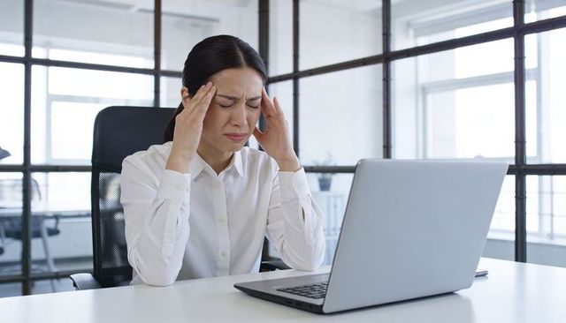Stressed asian businesswoman pressing temples at laptop in bright modern office workspace