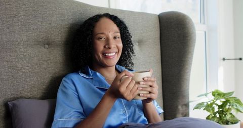 African american woman relaxing in satin pajamas sipping morning coffee in cozy bedroom