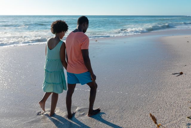 This image captures an African American couple walking hand in hand along a beach at sunset. Ideal for use in travel brochures, romantic getaway promotions, lifestyle blogs, and advertisements focusing on relationships, leisure, and summer vacations.