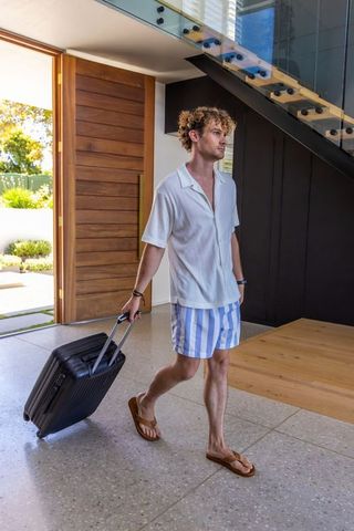 Young man walking through modern foyer pulling suitcase toward open pivot door