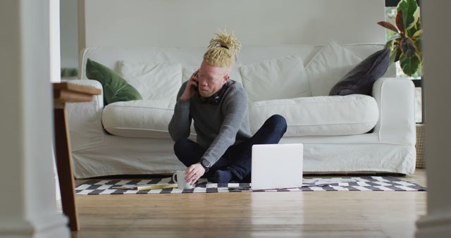 Young man with blonde dreadlocks working from home, sitting on the floor near a white couch. He is using a laptop and holding a cup of coffee while talking on the phone. This scene illustrates modern remote work, making it ideal for articles or ads on freelancing, technology, and work-life balance.
