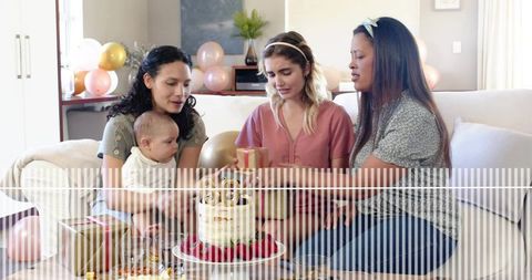Women celebrating birthday with baby, cake and gifts on cozy sofa
