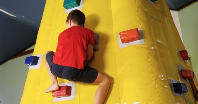A young boy wearing a red shirt and blue shorts climbs on an inflatable indoor climbing wall. The wall is bright yellow with multiple hand holds of different colors (red, blue, and gray). This image can be used to illustrate children's activities, indoor playgrounds, physical fitness, and sports training for kids.
