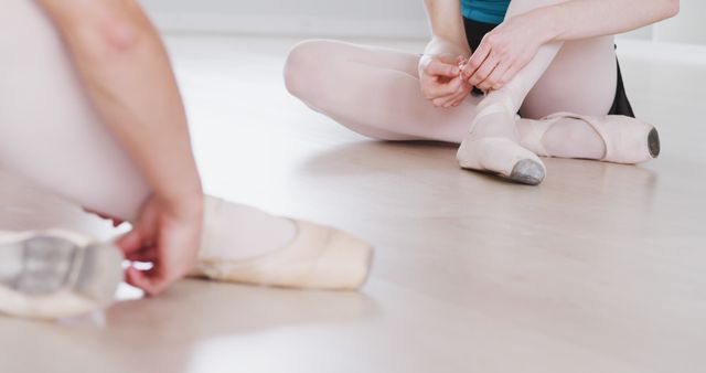 Two caucasian female ballet dancers sitting on floor tying pointe shoes at dance studio. Preparation, dance, ballet, practice and training, unaltered.