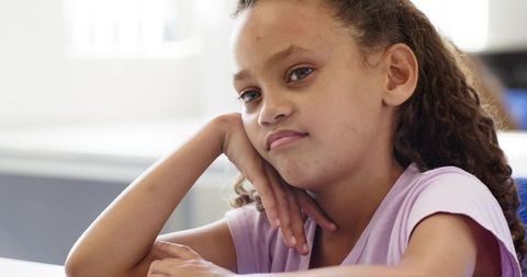 Thoughtful tween girl resting chin on hand by window, classroom portrait with curly hair