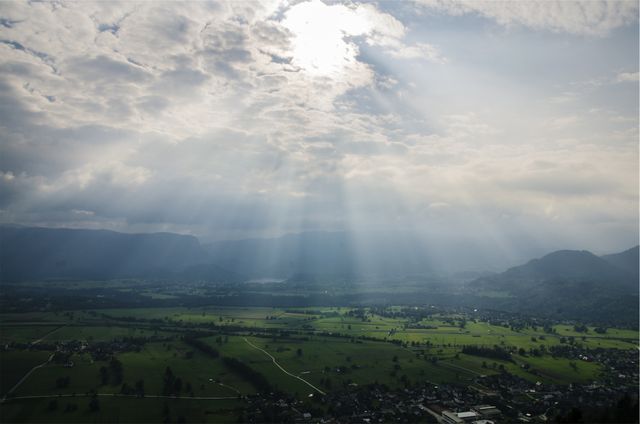 Sunlight is cutting through a cloudy sky, casting dramatic rays over a scenic valley. The landscape features green fields and scattered small villages, with mountains in the background. This image is ideal for illustrating concepts of hope, tranquility, or nature's beauty. Perfect for use in travel guides, nature blogs, inspirational content, and background images for various designs.