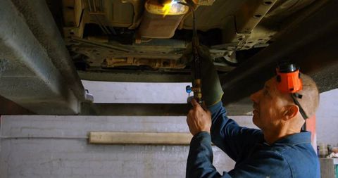 Mechanic torching vehicle undercarriage on lift wearing earmuffs and gloves, workshop