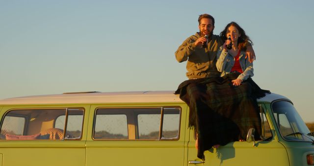 Couple sitting on roof of vintage van during sunset, covered in blankets and enjoying drinks. Perfect for depicting themes of love, travel, adventure, leisure, and relaxation. Ideal use for travel blogs, adventure magazines, advertisements for outdoor products, and lifestyle websites highlighting cozy moments and van life.