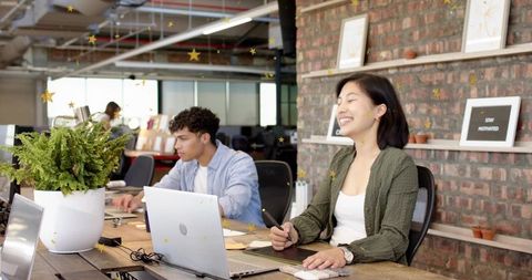 Smiling asian woman drawing on graphics tablet at modern coworking desk with laptops