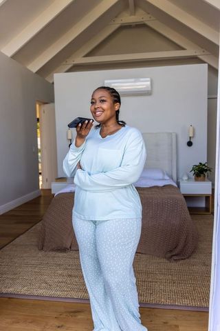 African american woman in pale blue pajamas speaking into smartphone in sunlit bedroom