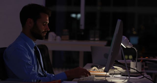 Young businessman working late hours at his desk in a dimly lit office. He appears to be focused on his computer, likely handling important tasks or meeting a deadline. Perfect for depicting dedication, corporate lifestyle, night shift productivity, and professional commitment in modern work environments.