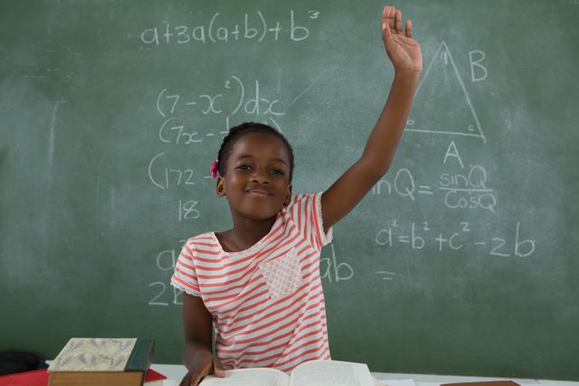 Young schoolgirl eagerly raising hand while sitting in classroom with chalkboard filled with mathematical equations. Ideal for educational content, school promotions, academic articles, and learning resources.