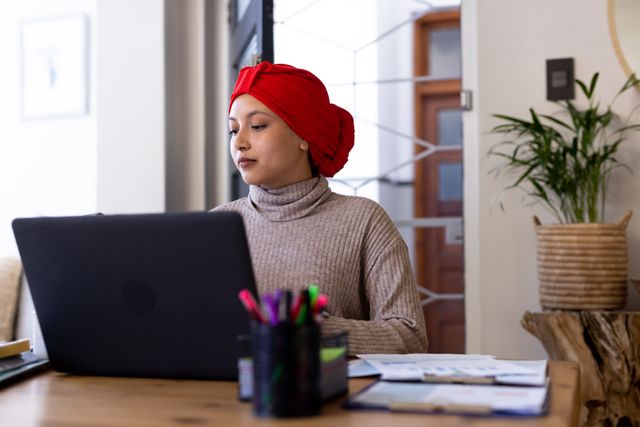 Serious biracial woman in hijab sitting at desk with paperwork, using laptop at home, copy space. Working from home, communication, inclusivity and lifestyle.