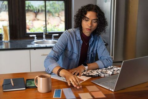 Non-binary designer arranging fabric and paint swatches at home kitchen workspace