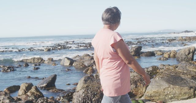 Senior woman is exploring a rocky beach during daytime. Theocean is calm in the background, and she is facing it while appearing to be looking at the water. She is wearing casual clothes and seems to be enjoying the natural surroundings. This can be used for depicting themes like outdoor activities for seniors, enjoying nature, independence, travels, and healthy lifestyle for the elderly.