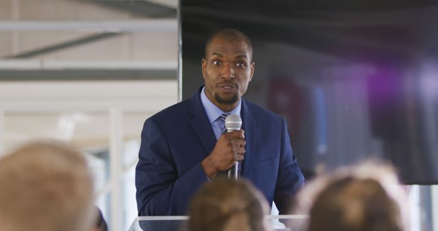Front view close up of a young African American businessman standing at a lectern using a microphone to address the audience at a business convention. The backs of the heads of the audience visible in the foregound