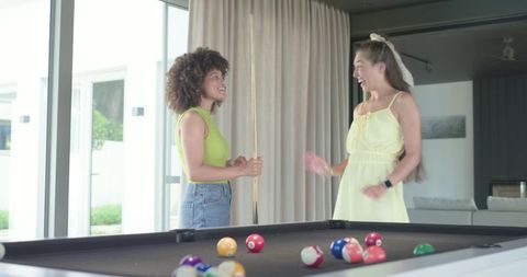 Diverse female friends laughing and playing pool in modern sunlit living room