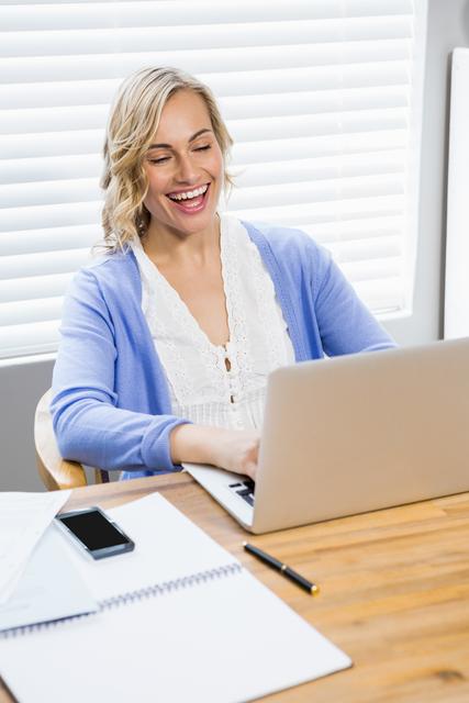 Woman seated at desk at home, using laptop and smiling. White blinds in background, wooden table with notebook, papers, smartphone, and pen. Perfect for illustrating remote work, home office setups, work-life balance, productivity, and technology use.