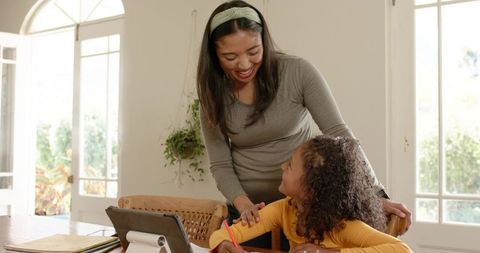 Smiling mother helping daughter with homework at sunlit kitchen table