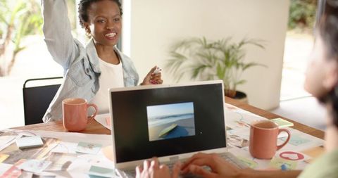 Diverse team brainstorming with laptop and sketches, woman raising hand in denim and notes