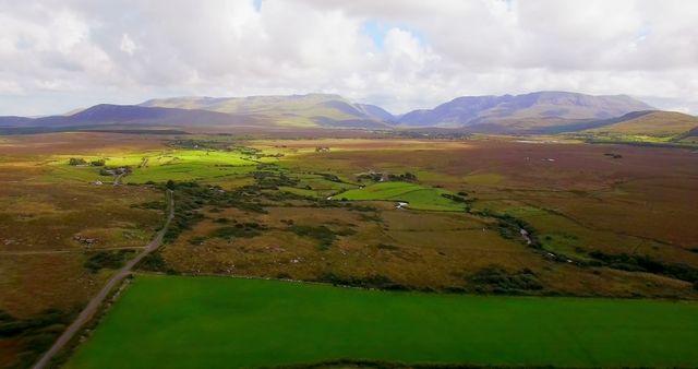 Aerial view of the Irish countryside featuring vast green fields, rolling hills, and a partly cloudy sky. The scenic beauty showcases a rural landscape with winding roads, scattered buildings, and mountains in the background. Ideal for use in travel magazines, nature documentaries, environmental blogs, or promotional material about Ireland's scenic landscapes.