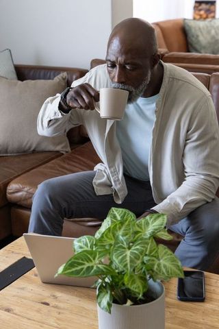 Mature african american man sipping coffee while checking laptop on living room sofa
