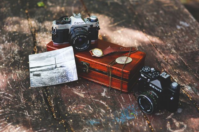 This image depicts two vintage cameras on a weathered wooden table next to an old black-and-white photograph and a small wooden box with a leather strap. Ideal for articles on photography, nostalgic decor, or retro-themed projects, it can be used for blog posts about film photography and vintage equipment or marketing materials for antique shops and vintage fairs.