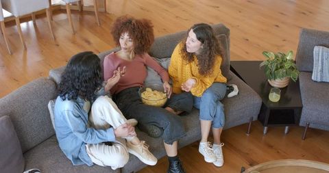 Multiracial women chatting on gray sofa sharing snacks and laughing in cozy living room