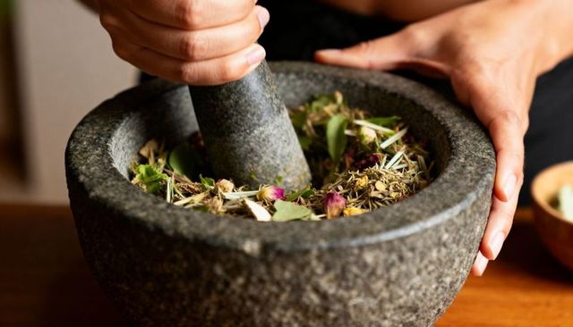 Grinding herbs in stone mortar with pestle and dried rosebuds on wooden counter