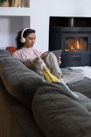 Non-binary person relaxing on gray sectional wearing headphones and using tablet by fireplace