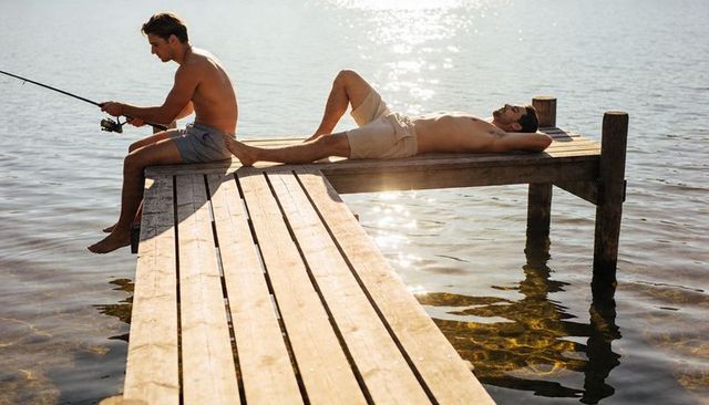Two men fishing and sunbathing on weathered wooden dock at golden lake sunset
