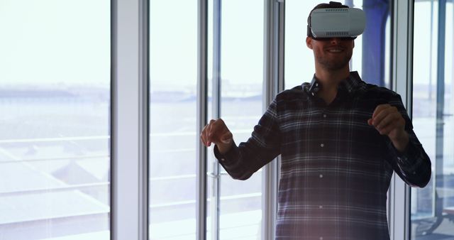 A man wearing a virtual reality headset indoors, standing near large windows. He appears to be enjoying an immersive experience as he interacts with the VR environment. This can be used in articles or advertisements about emerging technologies, virtual reality gaming, or innovation in entertainment.