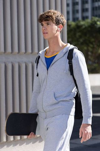 Teen walking with skateboard wearing grey hoodie and backpack along ribbed urban wall
