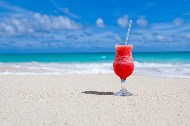 A vibrant red cocktail with a straw, standing on a sandy beach with turquoise ocean and clear blue sky in the background. This image is perfect for travel advertisements, vacation promotions, tropical holiday brochures, summer drink menus, and marketing materials for beach resorts.