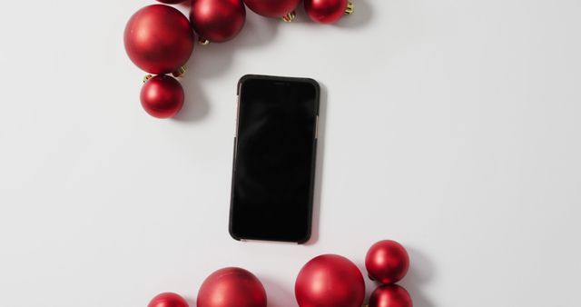 This image displays a smartphone placed among red Christmas ornaments on a white background, creating a festive and modern composition. Use this image to promote holiday sales for technology and mobile devices, seasonal campaigns for communication services, or to add a festive touch to tech-related content during the holiday season.