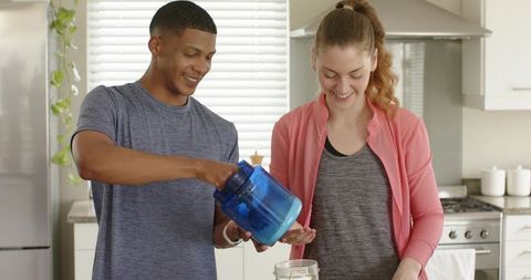 Diverse couple preparing supplement powder in bright modern kitchen for fitness