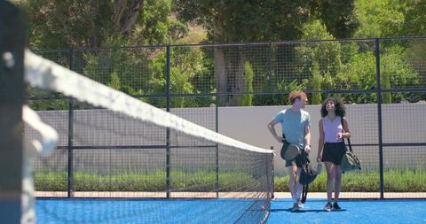 Young athletes walking on bright blue outdoor tennis court carrying rackets and sports bags