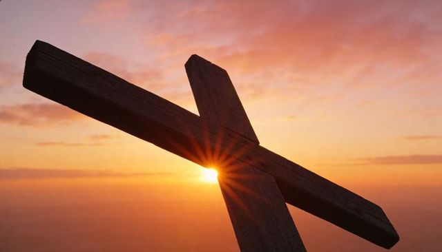 Rough wooden cross silhouetting against sunrise sky with starburst over horizon