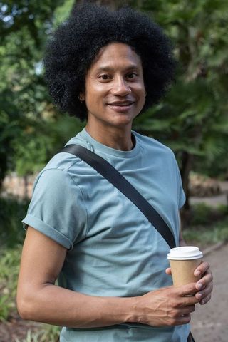 Smiling young man holding takeaway coffee with crossbody bag walking in leafy urban park