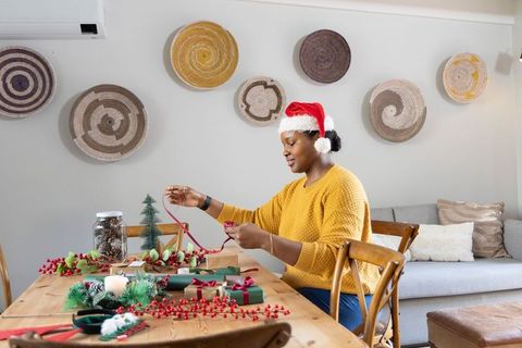 African american woman wearing santa hat tying ribbon wrapping gifts at dining table